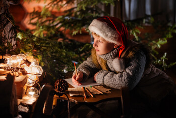 Merry Christmas and happy holidays! Little boy in winter clothes is writing letter to Santa Claus at night near the Christmas tree indoors.