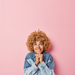 Dreamy curly haired European woman bites lips keeps hands under chin focused above dressed in denim jacket thinks about something isolated over pink background empty space for your promotion