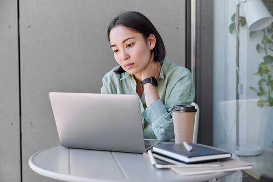 Asian female graphic designer watches tutorial about creative ideas focused at laptop screen involved in working process poses at table with notepads and takeaway coffee against cozy interior