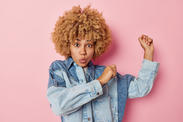 Carefree curly haired young European woman dances and raises arms up moves with rhythm of music wears denim jacket looks surprisingly at camera isolated over pink background foolishes around