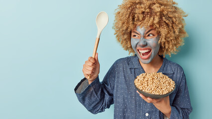 Angry curly haired woman yells with annoynace at loud neighbours holds bowl of cornflakes and spoon...