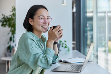 Skilled smiling freelancer sends file to customer via laptop computer takes note in notepad drinks tea from mug has dreamy cheerful expression enjoys coffee break. Millennial student studies distantly - obrazy, fototapety, plakaty