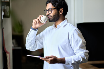 Smiling rich wealthy successful indian business man executive drinking morning coffee enjoying success at work having break starting day in office thinking af daily plans, looking through window.
