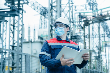 A masked power engineer during a pandemic inspects the modern equipment of an electrical substation...