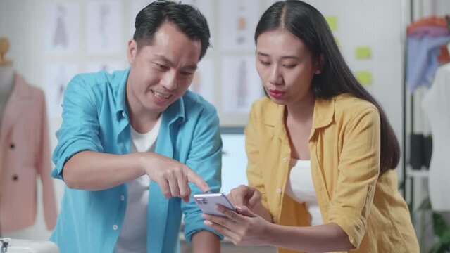 Asian Male And Female Designers With Sewing Machine Comparing Clothes Drawing Pictures On The Table To The Pictures On Smartphone While Designing Clothes In The Studio
