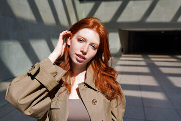 Teen stylish cool redhead fashion girl model standing on urban walls background looking at camera. Portrait of beautiful teenage girl with red hair lit with sun light outdoor.