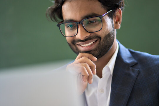 Happy arabic indian young business man executive professional financial manager, office employee wearing suit and glasses working on laptop looking at computer checking online data tech management.