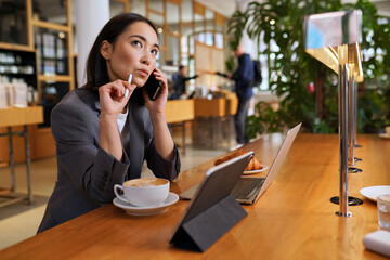 Doubtful Asian young adult business woman talking on cell phone making call working looking away thinking of solution sitting in cafe, considering offer, making decision or feeling doubt.