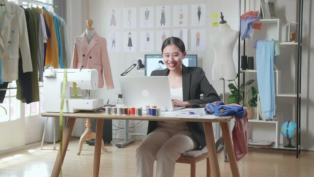 Smiling Asian Female Designer In Business Suit Working On A Laptop While Designing Clothes In The Studio
