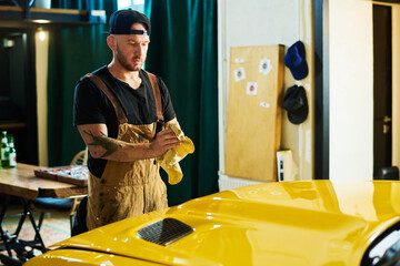 Fototapeta na wymiar Young repairman in workwear rubbing dirty hands with duster over hood of yellow electric car while standing in front of it in garage