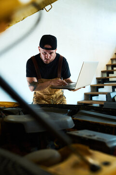 Young man bending forwards over open hood and using laptop while carrying out technical diagnostics of new installed equipment
