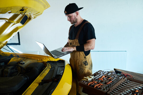 Young maintenance service worker with laptop standing in front of car with open hood and carrying out technical diagnostics