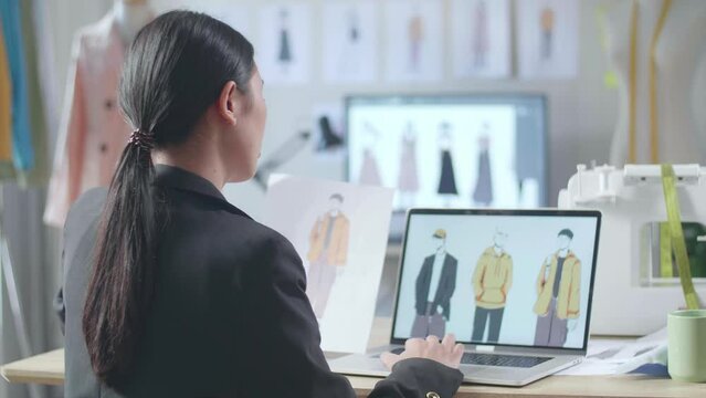 Back View Of Asian Female Designer In Business Suit Looking At The Paper In Hand And Comparing It To The Pictures On A Laptop While Designing Clothes In The Studio

