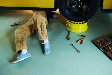 Legs of young repairman or technician in overalls lying under yellow electric car on the floor of...
