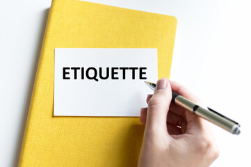 A white business card with ETIQUETTE text on a billboard on a white background next to the hand of a girl with a pen writes text