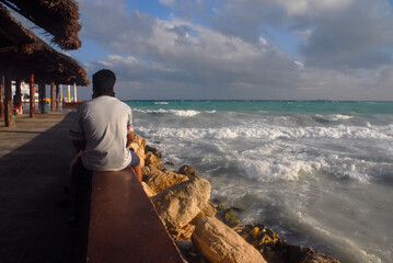 A rear view shot of a man sitting on a parapet as he watches the rough Caribbean Sea, and waves crashing on rocks in Mexico: Selective focus.