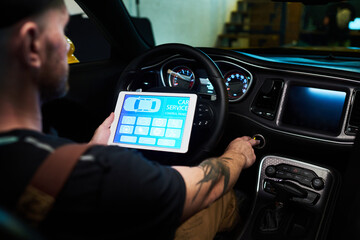 Young technician with digital tablet pressing button on instrument panel or dashboard of car while sitting in front of steering wheel