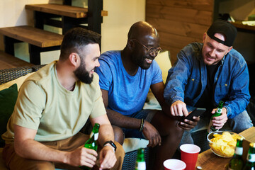 Cheerful buddies discussing photos in smartphone of African American guy while sitting by table with bottles of beer and snacks