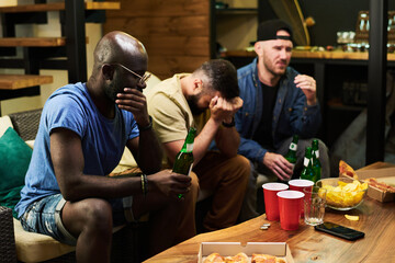 Group of upset young buddies expressing sadness about their favorite football team while sitting in front of tv set and watching broadcast