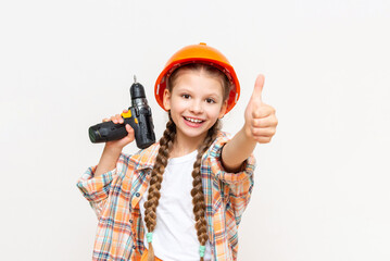 A little girl in a construction helmet and a drill in her hands gives a thumbs up and smiles on a white isolated background.