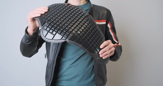 Biker in a leather black sports motorcycle jacket, holds a plastic back protection in his hands, demonstrates it, squeezes it. Front view, close-up, indoors.