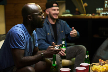 African American guy with controller looking at tv screen during video game while sitting against his buddy commenting actions