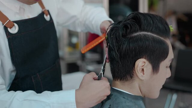 Asian professional male using scissors cutting customer hair in salon. Attractive hairdresser man using scissors cut hair young men and looking at mirror with smiling. Beauty salon business concept.