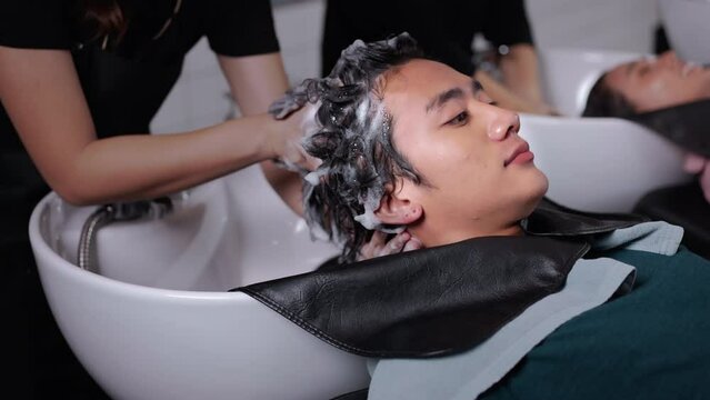 Asian attractive young male get hair washed by hairdresser in salon. Professional hair stylist woman give service and hair wash and massage to handsome man customer lying down on salon washing bed.