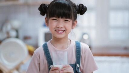 Portrait of Asian little kid holding cup of milk in kitchen in house. Young preschool child girl or daughter stay home with smiling face, feel happy enjoy drinking milk and then looking at camera.