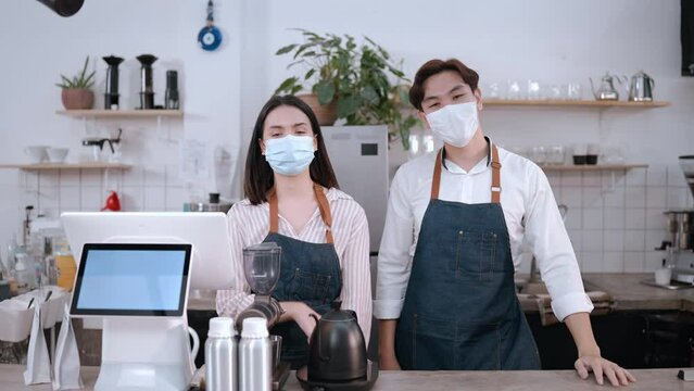 Portrait of businessman and woman wearing face mask standing in cafe. Attractive young barista business couple wear protective mask due pandemic stand with confident and crossing arm at coffee shop. 