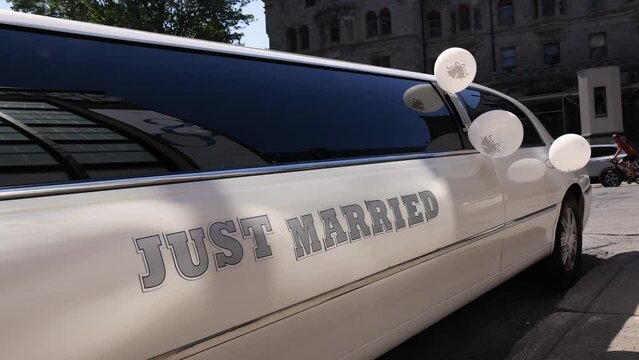 Side angle view of a white stretch limousine with the words just married and celebration balloons attached to windows parked near a wedding venue.
