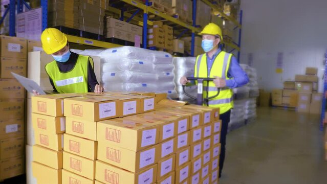 Retail worker in uniform is moving cardboard boxes by using hand pallet truck and stop to check quality amount of the product by his manager use with digital laptop device in the warehouse.