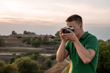 Young man holding retro camera and taking pictures. He is wearing green shirt. Fortress and sunset are in background.