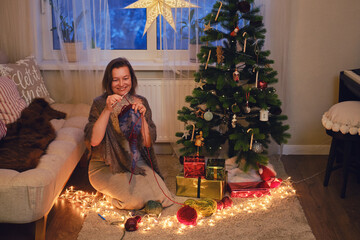 An adult woman knits clothes by the Christmas tree in the evening living room. Hobbies and...