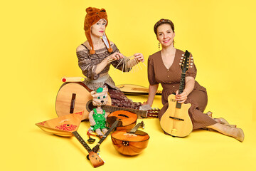 Women musicians in dresses with musical instruments on a yellow studio background. Happy artists...