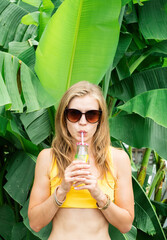 young caucasian woman in yellow swuimsuit drinking beverage standing in tropical palm leaves
