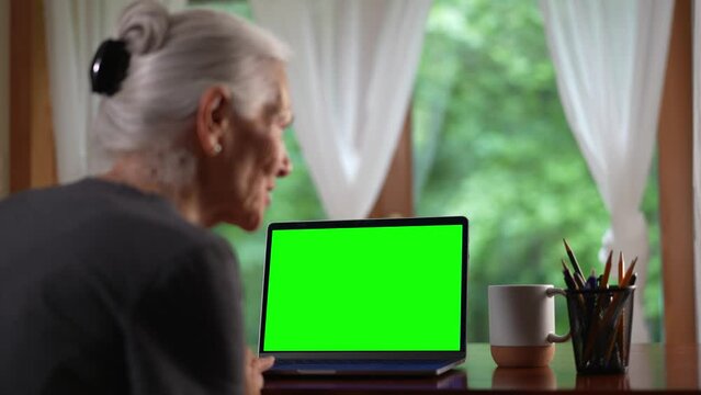 Over shoulder view of senior elderly woman making video call on laptop green screen talking and listening during video chat with friends or colleagues.