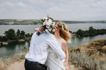 The bride and groom are hugging outdoors against the backdrop of the river and are closed with a...