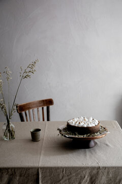 Still life table setting with a swiss meringue pie on the table, a wooden chair, a ceramic cup, and a dried oats bouquet 