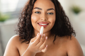 Portrait of black body positive lady applying lip balm moisturizing skin, looking at camera - obrazy, fototapety, plakaty