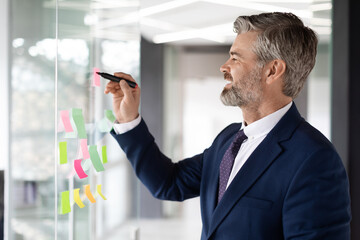 Mature Male Entrepreneur Writing On Sticky Notes On Glass Wall At Office