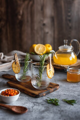 Empty glass mugs decorated with rosemary for serving sea buckthorn tea.  A warming winter drink. Vitamin tea. Sea buckthorn tea with honey, lemon and ginger on a gray background.