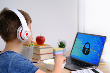 smart child in headphones, boy 7-8 years, primary school student writes in front of open laptop,...