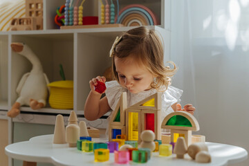 Obraz na płótnie Canvas A little girl playing with wooden blocks on the table in playroom. Educational game for baby and toddler in modern nursery. The kid builds a tower from wooden rainbow stacking blocks.
