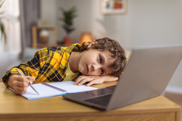 Boring upset little boy lying on hand and writing in copybook, watching lesson online on laptop, empty space © Prostock-studio