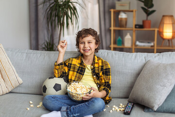 Happy little boy having fun, watching football on tv and eating popcorn, sitting on messy sofa and...