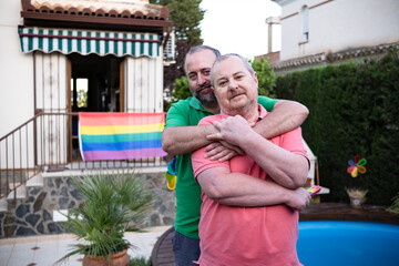 Middle-aged couple embracing in the garden of their home with an LGBT flag in the background.