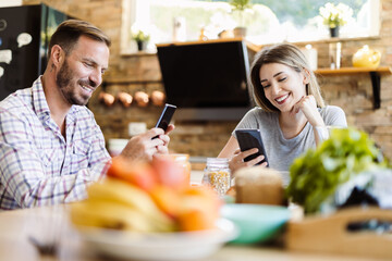 Young happy couple using mobile phones during breakfast time at home
