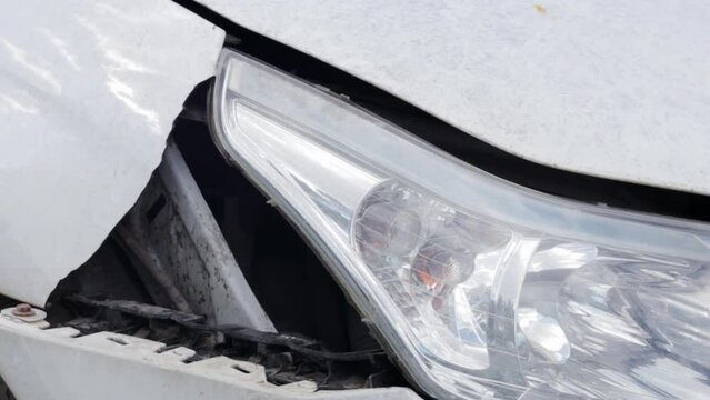 Accident on the street, damaged car after a collision in the city. Broken right headlight as a result of a collision. Broken white car after an accident. Damaged headlight, body, hood and bumper.