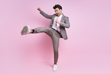 Excited young man in full suit throwing leg kick while standing against pink background